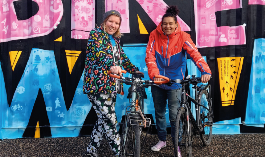 Two women holding hybrid bikes standing in front of a bright mural